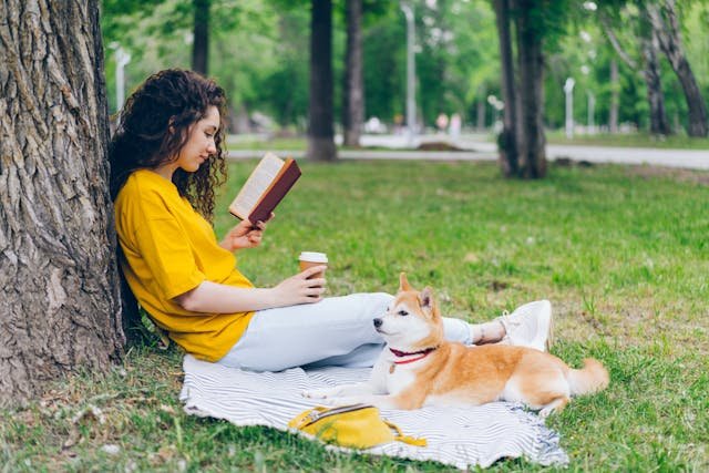 A girl sitting on a blanket under a tree with her dog while reading a book